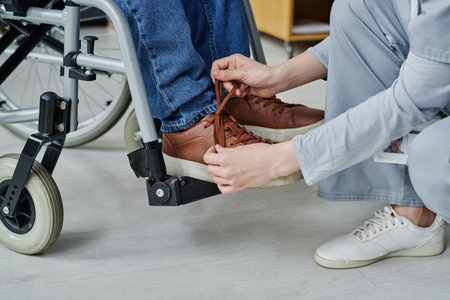 Close-up of young woman helping man with disability to tie shoelaces while he sitting in wheelchairの写真素材