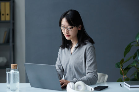 Asian young businesswoman in eyeglasses concentrating on her online work on laptop sitting at her...