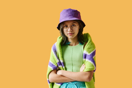 Portrait of teenage girl in bright hat and eyeglasses standing with her arms crossed and looking at camera against orange backgroundの写真素材