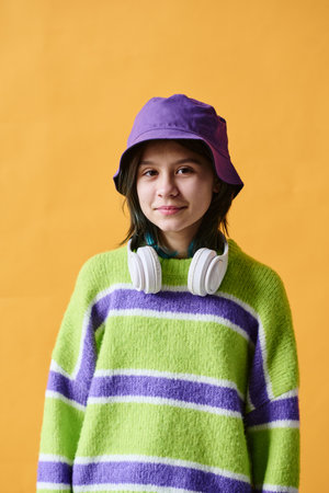 Portrait of teenage girl in bright hat and wireless headphones looking at camera standing against orange backgroundの写真素材