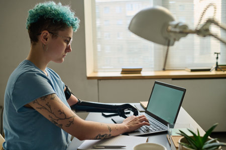 Young woman with prosthetic arm typing on laptop and working online at table in the roomの写真素材
