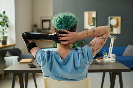 Rear view of young woman with prosthetic arm resting at table after online work in the roomの写真素材