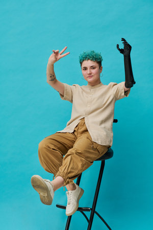 Vertical image of teenage girl with prosthetic arm sitting on chair against blue background