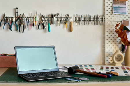 Laptop with black screen standing on workplace of leather worker with hammer, color swatches, handtools on wall and other suppliesの写真素材
