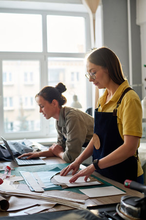 Side view of female tailors working in atelier or studio of fashion while one of them networking and the other going to circle sewing patternsの写真素材