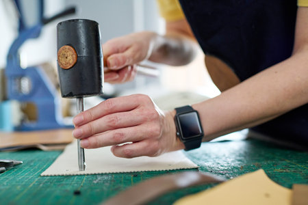 Professional female tanner keeping hammer on top of small steel shaft while pressing it down leather piece during work in craft shopの写真素材