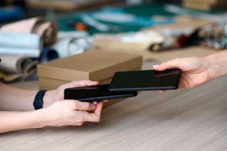 Close-up of hand of young female buyer holding smartphone over that of seller while paying for...
