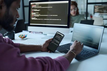 Young programmer sitting at his workplace and working with computer codes, he connecting smartphone with laptopの写真素材