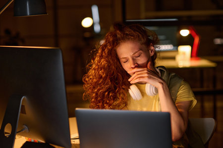 Young tired woman sitting at her workplace in front of computer and yawning, she working till late nightの写真素材
