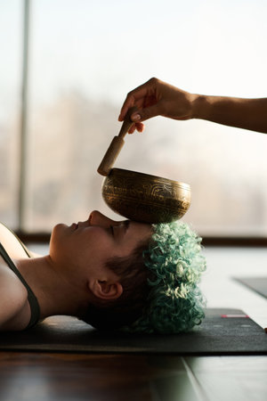 Vertical image of young girl lying on the floor on exercise mat and relaxing with singing bowl on her face during practiceの写真素材