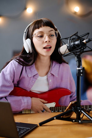 Vertical image of singer with guitar recording her song, singing in microphone at tableの写真素材