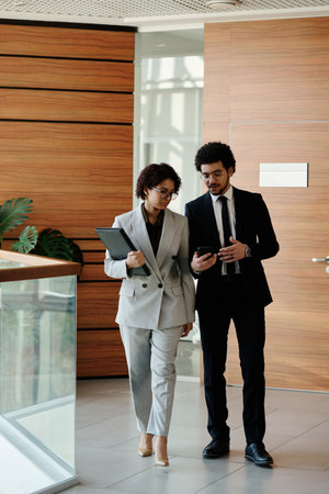 Two young colleagues talking to each other while walking along office corridorの写真素材
