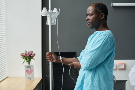 African American elderly patient standing with dropper in ward and looking through the window with pensive expressionの写真素材