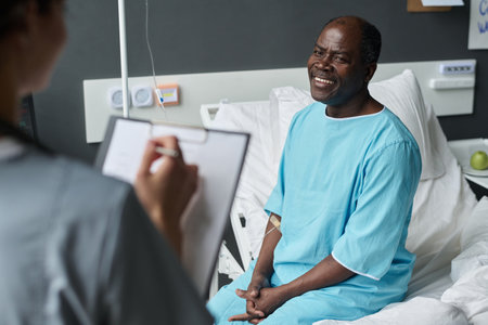 Smiling elderly patient sitting on bed and talking to nurse about his health condition while she making noted in medical cardの写真素材