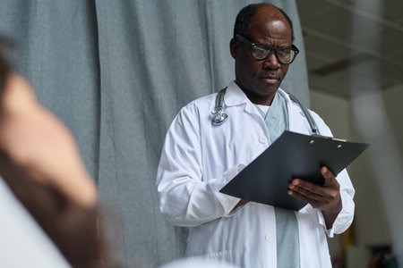 African American doctor in white coat writing prescription for patient while standing in ward