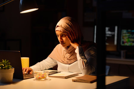 Woman in hijab working online on laptop till late evening at her workplace in officeの写真素材