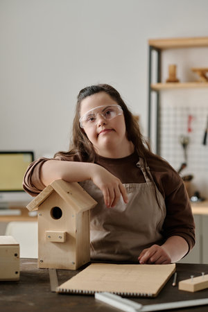 Vertical portrait of girl with down syndrome looking at camera while making house for birds in workshopの写真素材