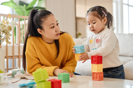 Asian young mother talking and playing with her little daughter in colorful blocks in the room