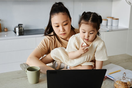 Asian young woman working online on laptop while sitting with her child at homeの写真素材