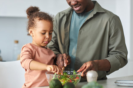 African American little girl helping her dad to cook vegetable salad in the kitchenの写真素材