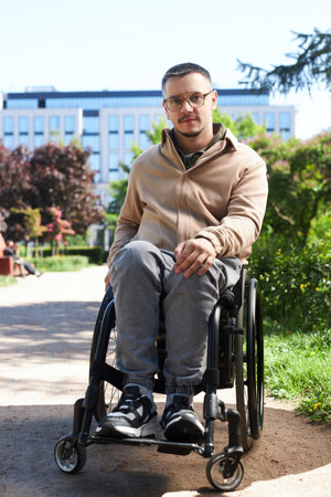 Vertical portrait of young man sitting in wheelchair and looking at camera during his walk in the city in summerの写真素材