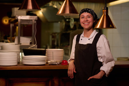 Young cheerful female worker of cafe or restaurant in uniform looking at camera while standing by counter with plates and waiting for guestsの写真素材