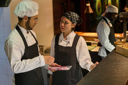 Young serious woman in uniform looking at male chef with meat fillet in container during discussion of new points of restaurant menuの写真素材