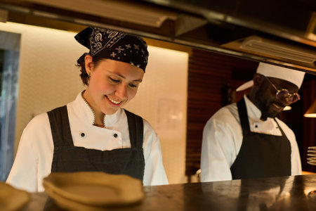 Happy young woman in uniform and her African American male colleague standing by counter and cooking food for guests of restaurant or cafeの写真素材