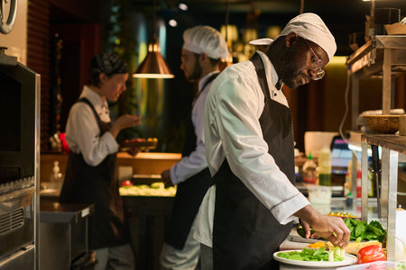 Young serious chef of restaurant mixing fresh chopped vegetables on round board while standing by workplace against two colleaguesの写真素材
