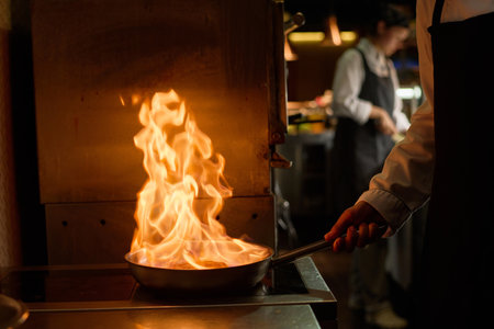 Close-up of young male chef holding frying pan with burning fire by handle while standing by stove in the kitchen and preparing new dishの写真素材