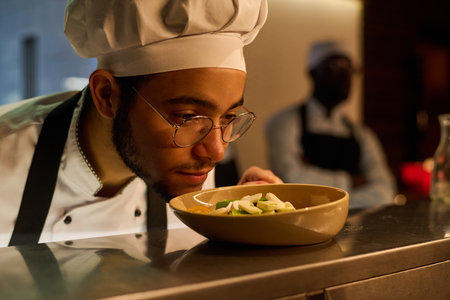 Face of young chef in uniform enjoying smell of fresh vegetarian salad in plate while bending over...