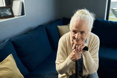Pensive old woman in beige casualwear leaning on walking cane while sitting on blue sofa in living room and thinking of her familyの写真素材
