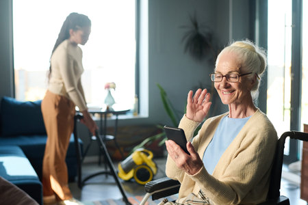 Happy senior woman with disability waving hand to someone on mobile phone screen while communicating with her family in video chatの写真素材