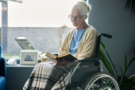 Senior woman with disability sitting in wheelchair by large window in living room and reading book...