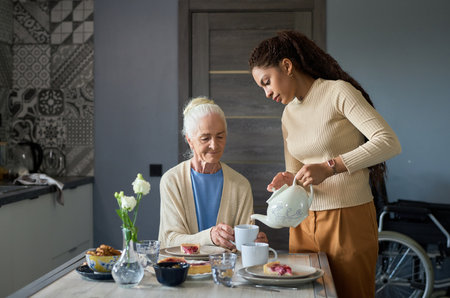 Cute teenage girl with porcelain teapot pouring tea into cup of her grandmother sitting by table in the kitchen during breakfastの写真素材