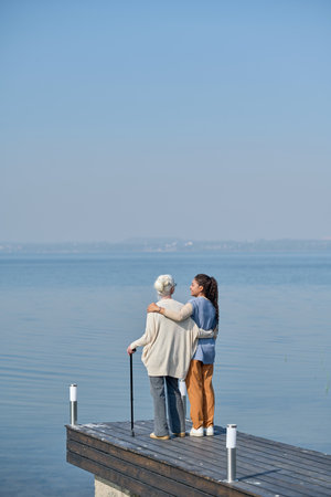 Rear view of granmother and granddaughter embracing each other while standing on pier and enjoying strolling together by watersideの写真素材