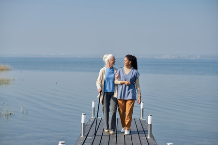 Long shot of aged woman and her granddaughter walking along pier against blue lake and sky and looking at one another while chattingの写真素材