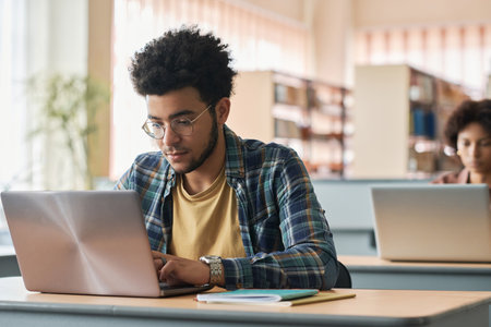 Young migrant sitting at desk and studying online with laptop with other students in classの写真素材