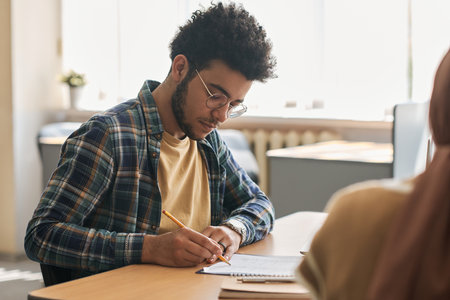 Migrant student sitting at table and writing exam with other students in classの写真素材