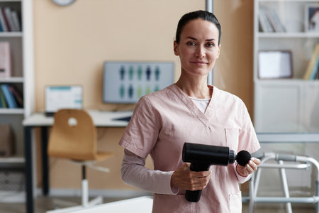 Portrait of physiotherapist in uniform using massager in her work, she standing in her office and...