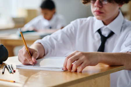 Schoolboy sitting at desk and making notes in his notebook during lessonの写真素材
