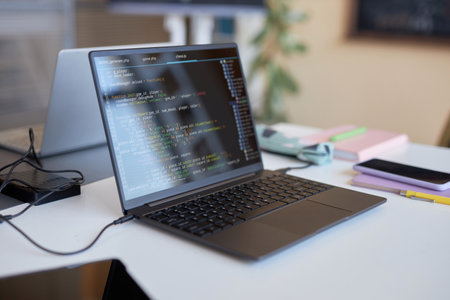 Laptop with codes on screen standing on desk of highschool student with smartphone, pen, and other...