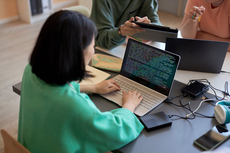 Young female student typing on laptop keyboard while decoding data on screen against two classmates...