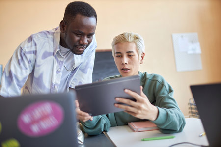 Confident Asian male student with tablet explaining his classmate data on screen while both...