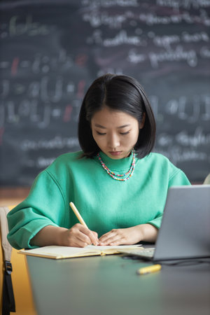 Serious Asian girl in casualwear making notes in copybook at lesson while sitting in front of laptop with blackboard with formulas on backgroundの写真素材