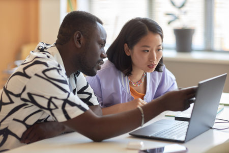 Confident African American guy pointing at online data on laptop screen while explaining Asian...