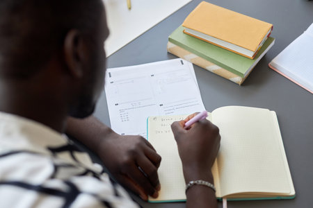 Close-up of young black man writing down formula or equation on page of copybook while sitting by desk and answering test questionsの写真素材