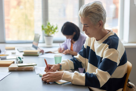 Side view of young male student in striped pullover making notes in copybook while sitting by desk...