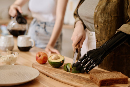Close-up of girl with prosthetic arm cutting vegetable for breakfast while her girlfriend pouring coffee in backgroundの写真素材