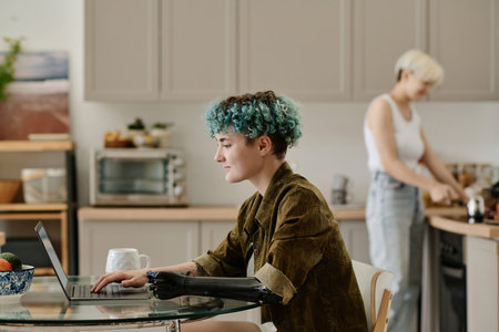 Girl with prosthetic arm working on laptop while sitting at table in the kitchenの写真素材